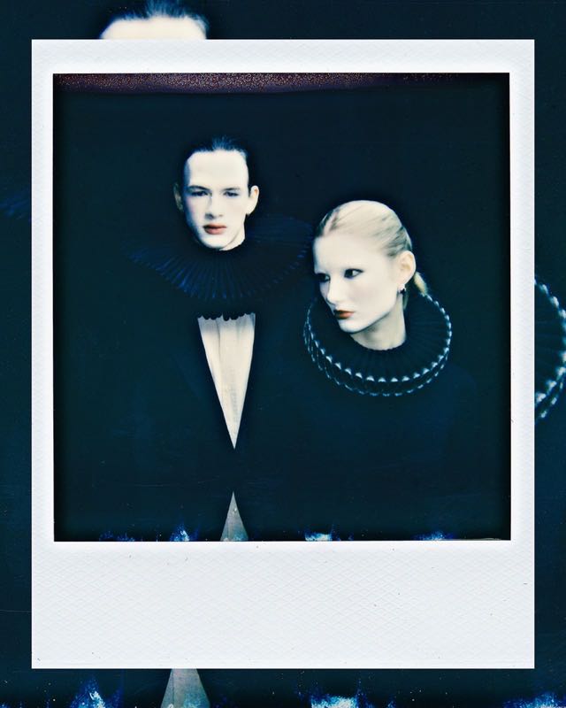 Polaroid mit zwei blass geschminkten Gesichtern vor dunklem Grund, blau getönt
