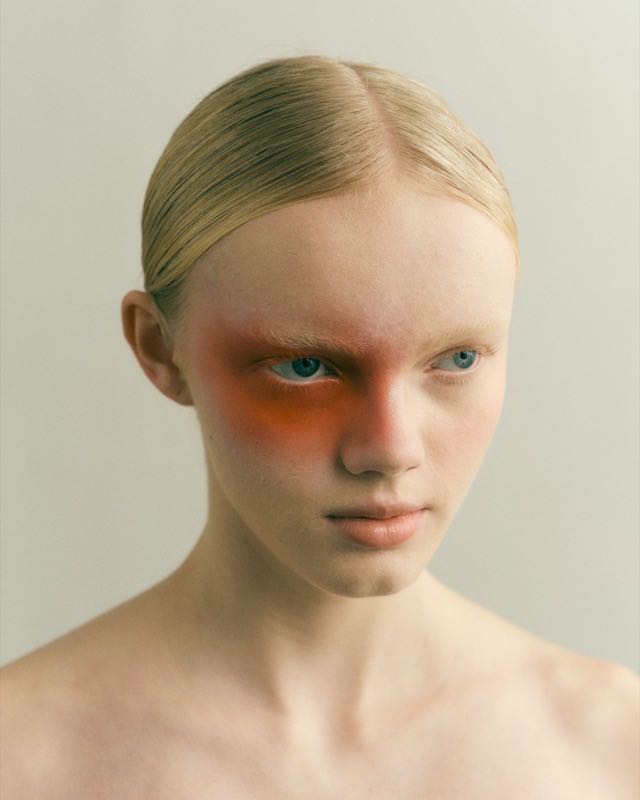 Porträt mit orangefarbenem Make-up um das linke Auge vor hellem Grund